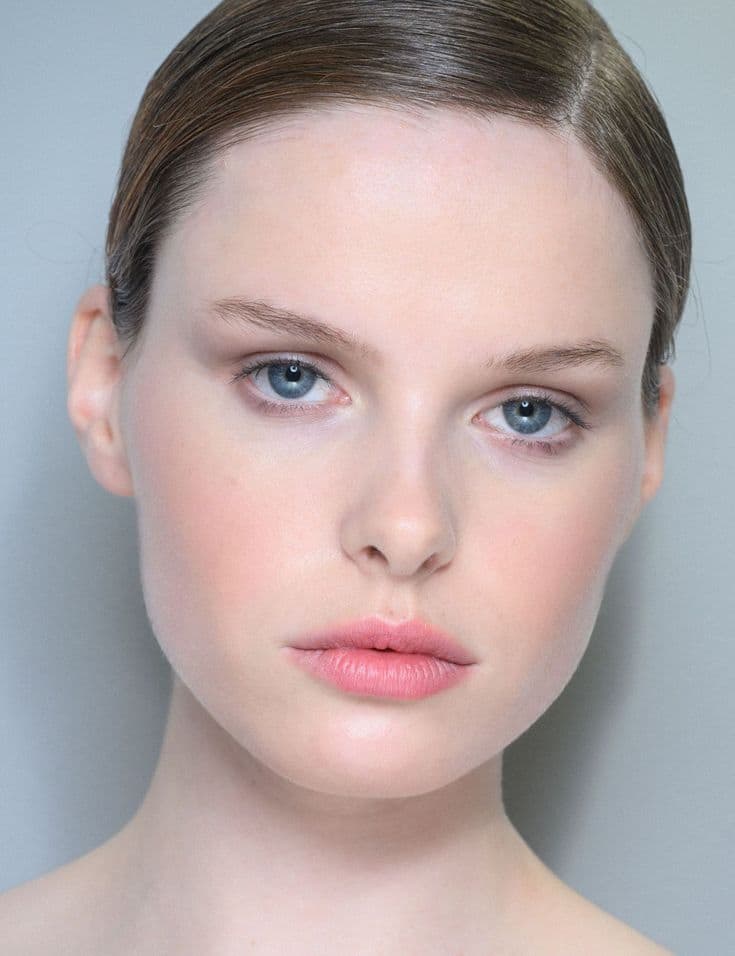 Primo piano di una modella con carnagione chiara e luminosa. Il trucco si basa su tonalità fredde e delicate: un blush rosa tenue sulle guance, labbra color rosa confetto opaco e un ombretto leggerissimo che risalta gli occhi azzurri. Lo sfondo è di un grigio freddo e minimale